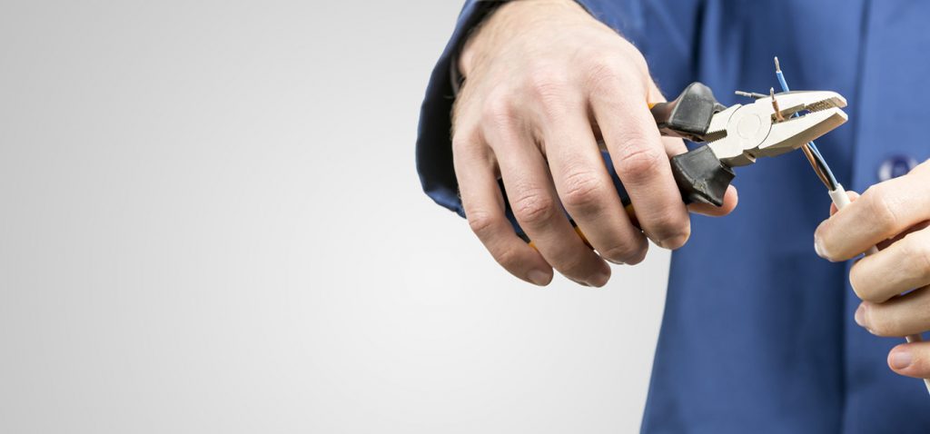 Workman or electrician repairing an electrical cable with a pair of pliers to restore supply to the house, close up view of his hands in blue overalls on grey with copyspace