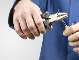 Workman or electrician repairing an electrical cable with a pair of pliers to restore supply to the house, close up view of his hands in blue overalls on grey with copyspace